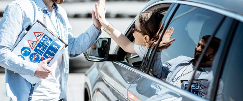 Moniteur d'auto-école et élève conductrice en voiture se tapent dans la main. Il tient un clipboard avec signalisation routière (vitesse, virage, interdictions).