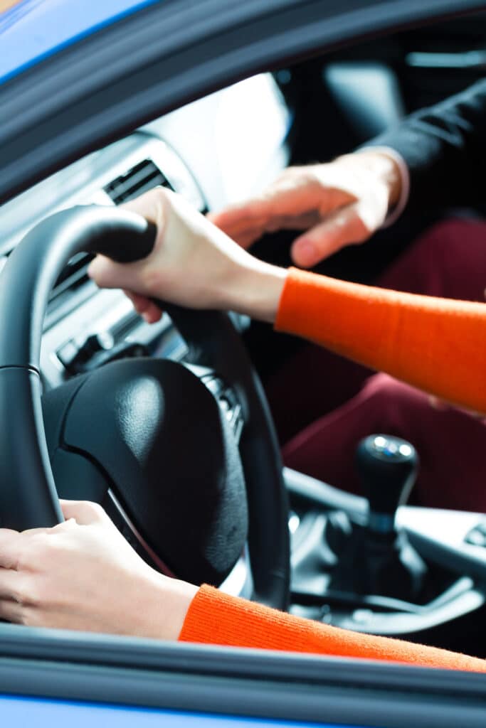 Mains d'un élève en pull orange sur un volant, guidées par un instructeur en costume noir, dans une voiture, pour un cours de conduite.